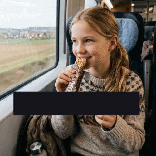 Kind sitzt am Zugfenster und isst einen Riegel, draußen zieht die Landschaft vorbei
