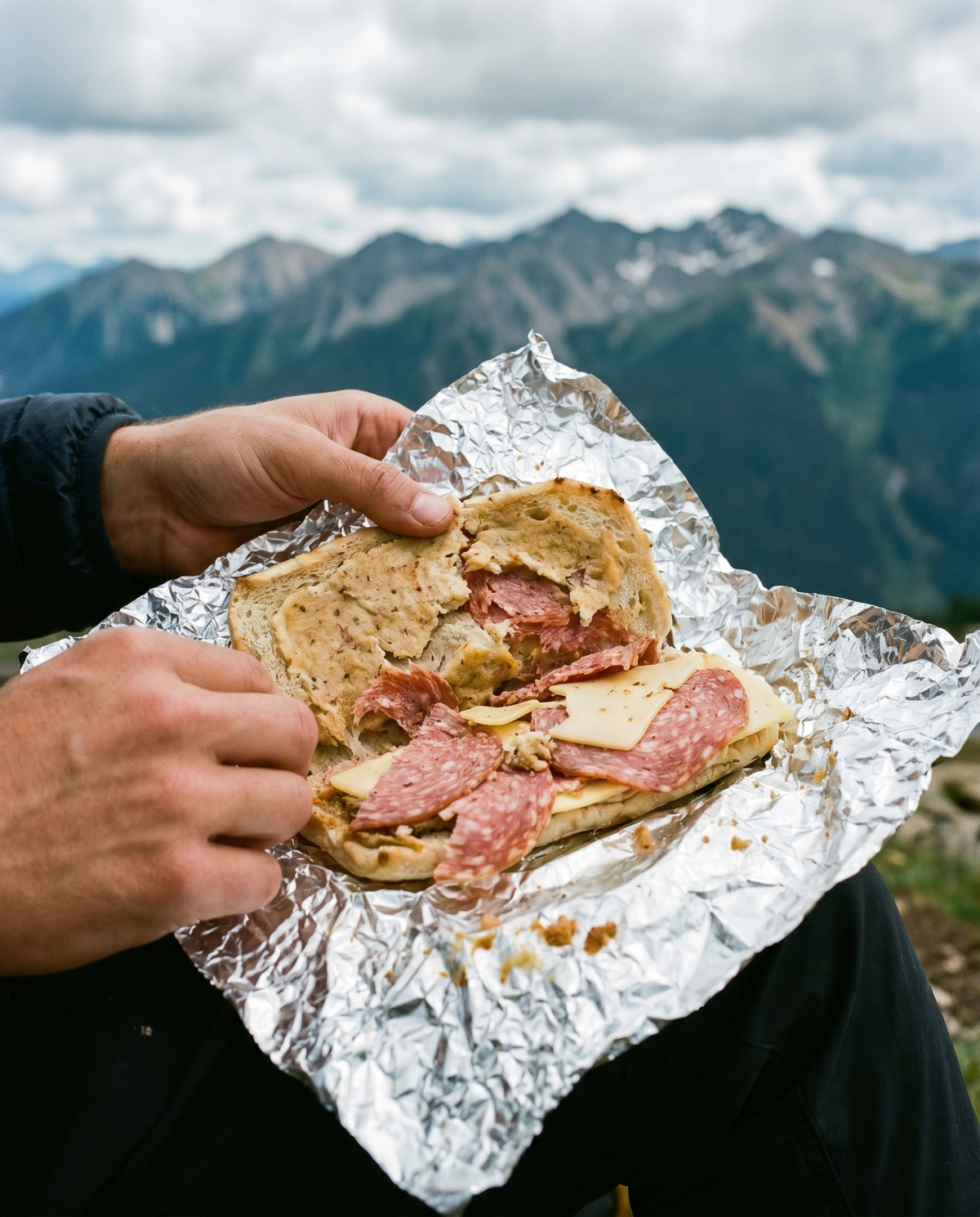 Hände halten ein in Alufolie eingeschlagenes Salamibrot vor einer Bergkulisse