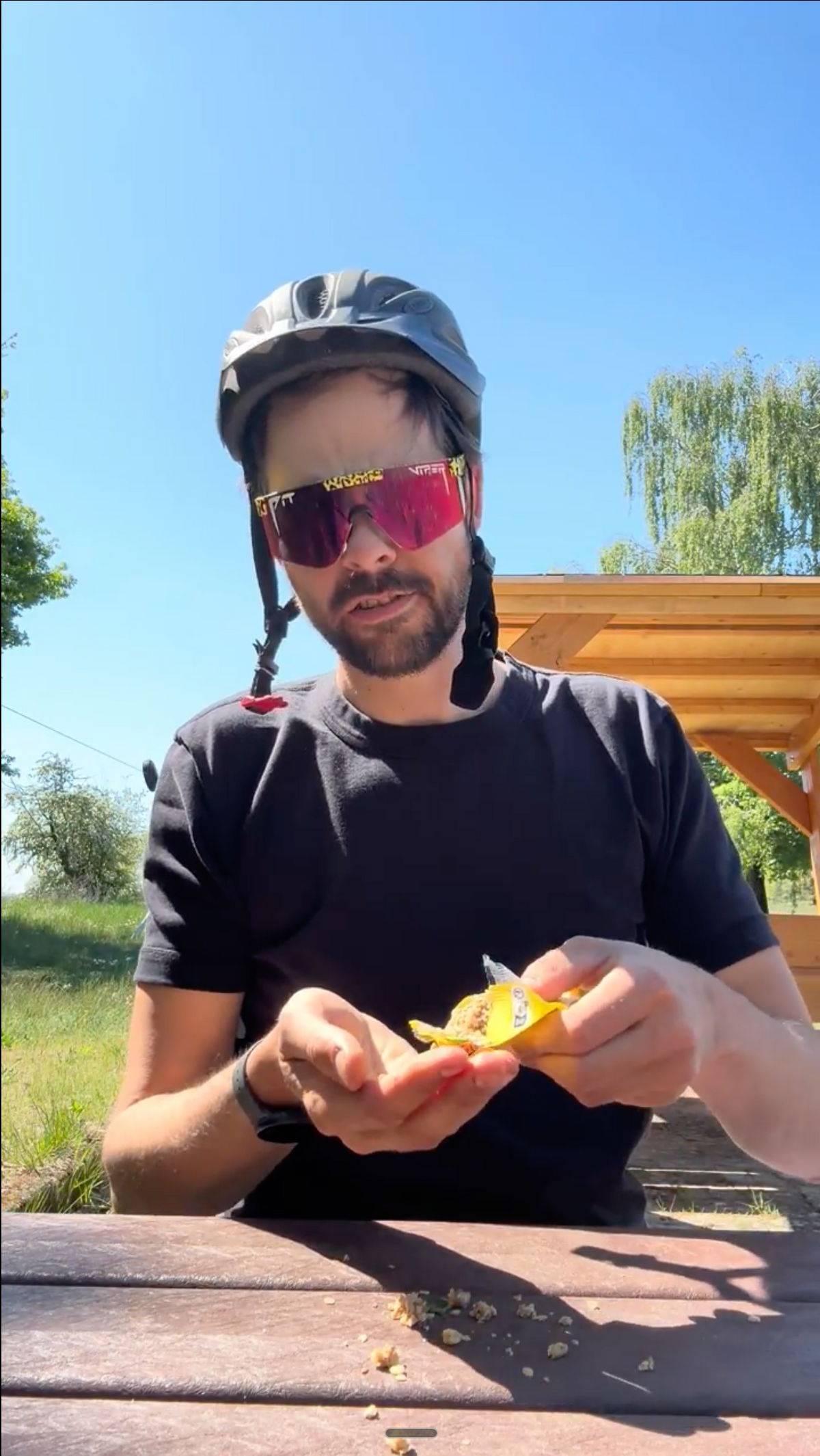 Person mit Fahrradhelm und Sportbrille sitzt an einem Holztisch im Grünen und packt einen Riegel aus