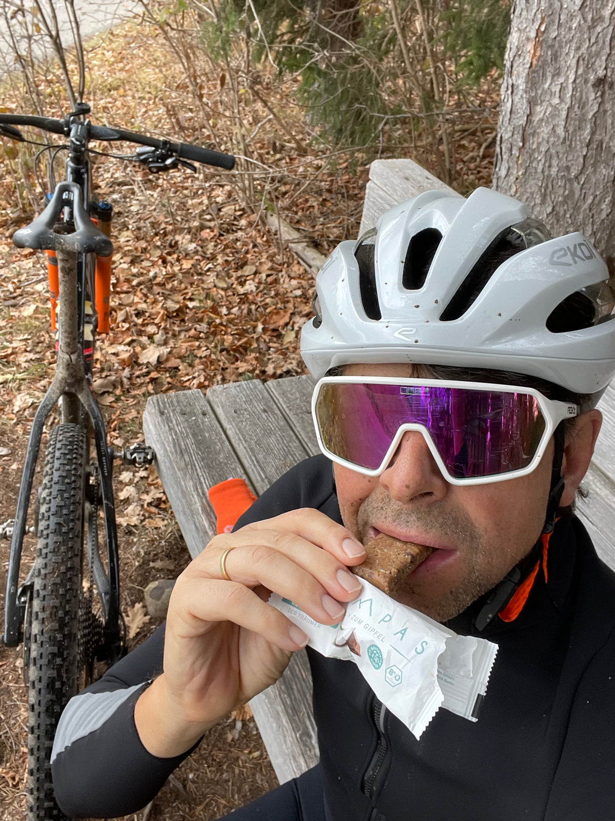 Pause am Waldrand: Person mit Fahrradhelm und Sportbrille isst einen TZAMPAS-Riegel, daneben lehnt ein Mountainbike