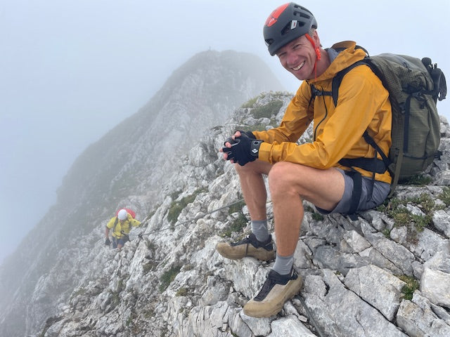 Person in gelber Jacke mit Kletterhelm sitzt lachend auf einem Felsgrat im Nebel