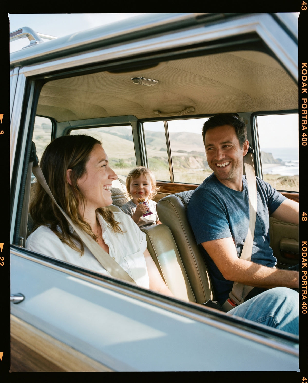 Familie im Auto: zwei Erwachsene vorn lachen, dahinter sitzt ein Kleinkind im Kindersitz, draußen Küstenlandschaft
