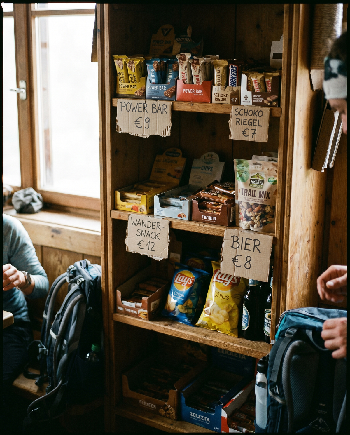 Verkaufsregal in einer Berghütte mit handgeschriebenen Preisschildern: Power Bar 9 Euro, Schoko Riegel 7 Euro, Wander-Snack 12 Euro, Bier 8 Euro